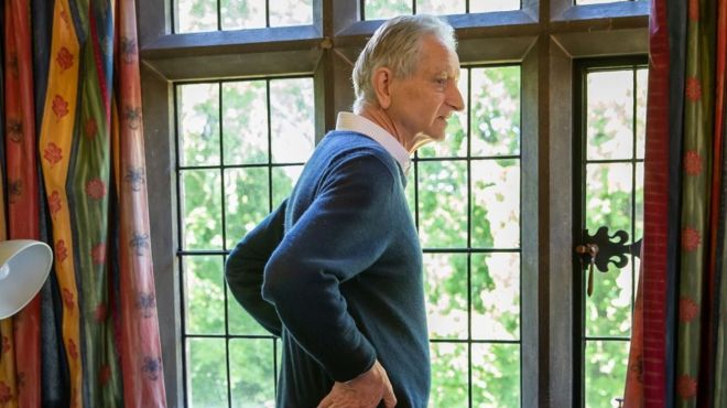 A man with grey hair and a blue jumper stands looking out of a stone window