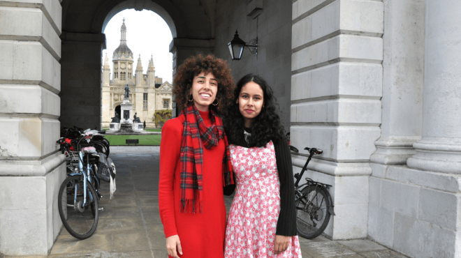 Two smiling women standing under a classical stone archway with the King's College gatehouse in the distance