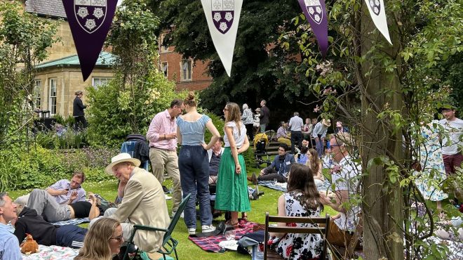 A group of people enjoy a relaxed picnic in a sunny garden decorated with purple and white bunting.