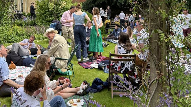 A group of people enjoy a relaxed picnic in a sunny garden decorated with purple and white bunting.