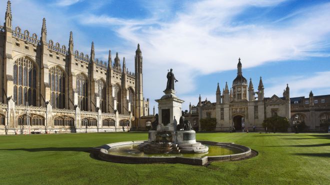 A large gothic style Chapel is in view to the left of an immaculate green lawn. In the middle there is an ornate water fountain with two figures to the side and a figure on top. In the background there is a gatehouse with spires. The sky is bright blue with gentle clouds.