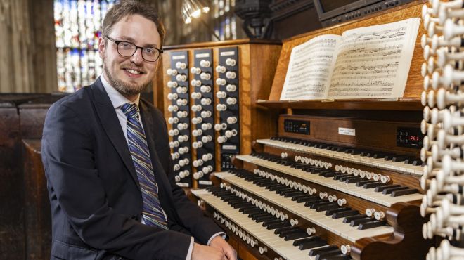 A white man with glasses wearing a suit sits to the left of an organ keyboard