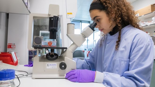 A young woman looks into a microscope wearing a lab coat and gloves