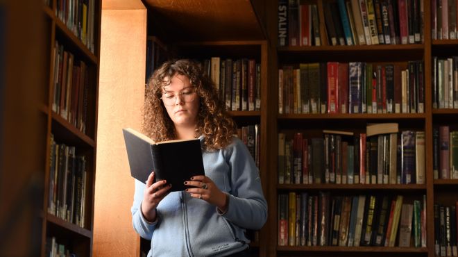 a woman stands by a window reading in a library