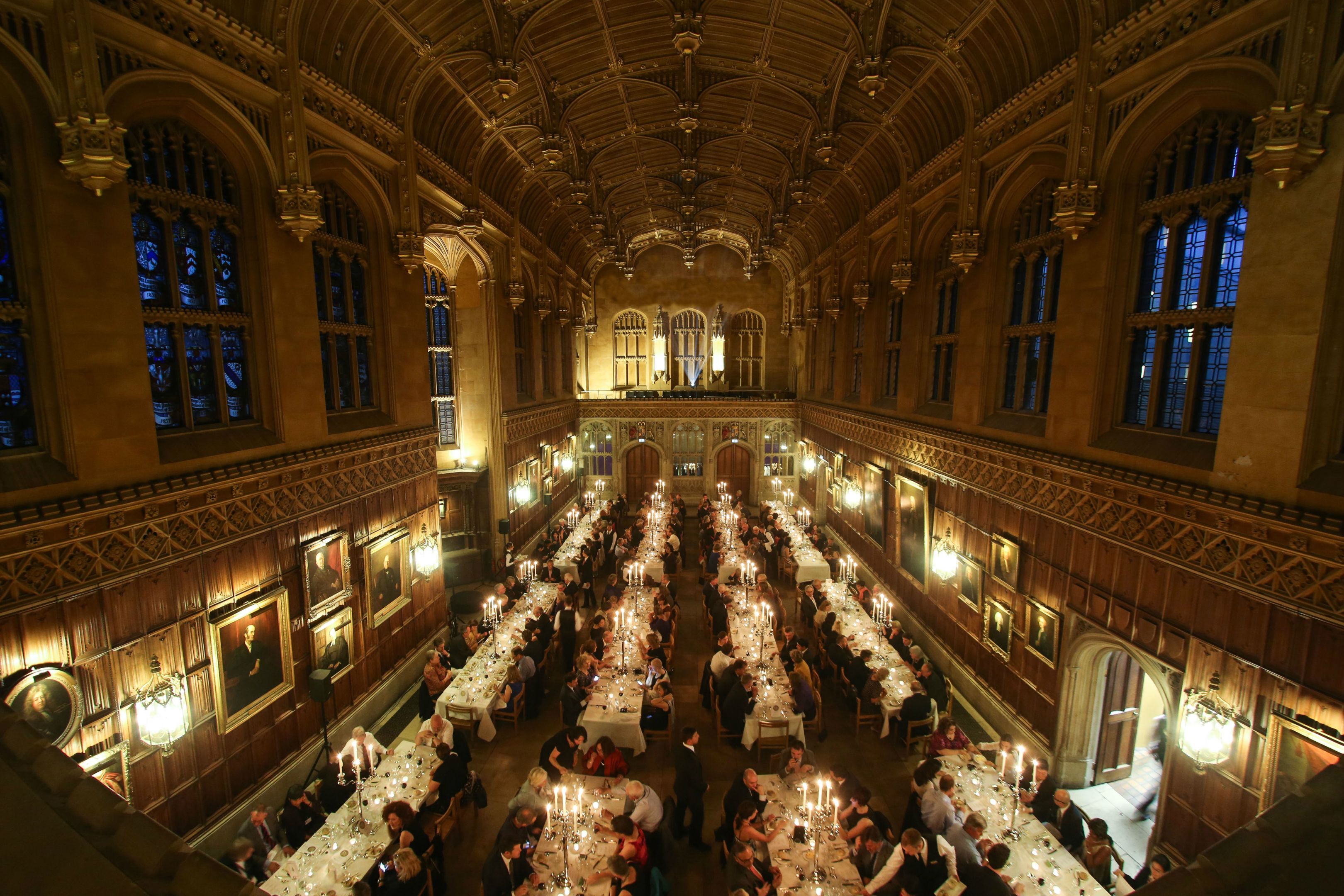 A candlelit dining hall wtih long tables and formal place settings