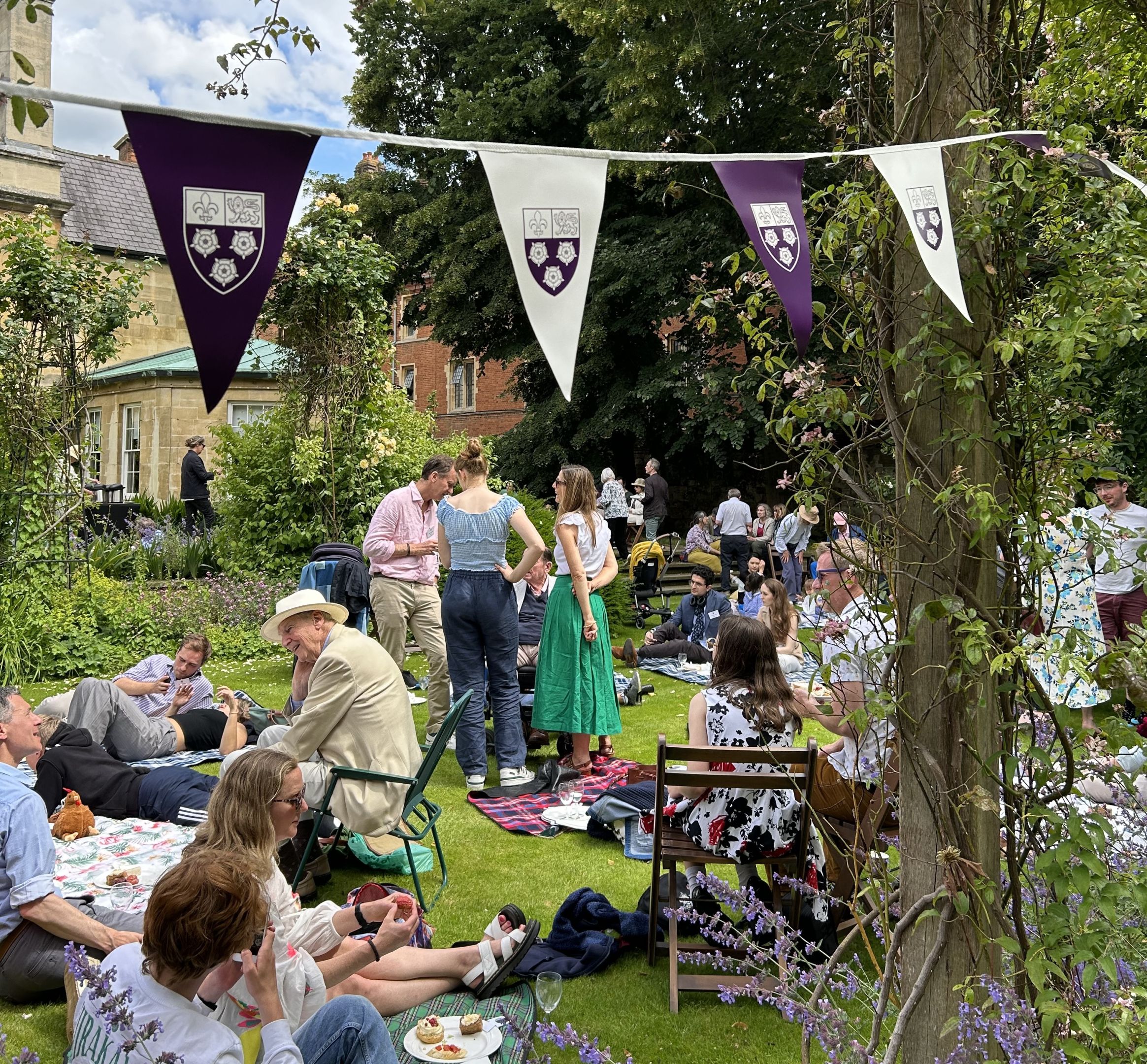 A group of people enjoy a relaxed picnic in a sunny garden decorated with purple and white bunting.