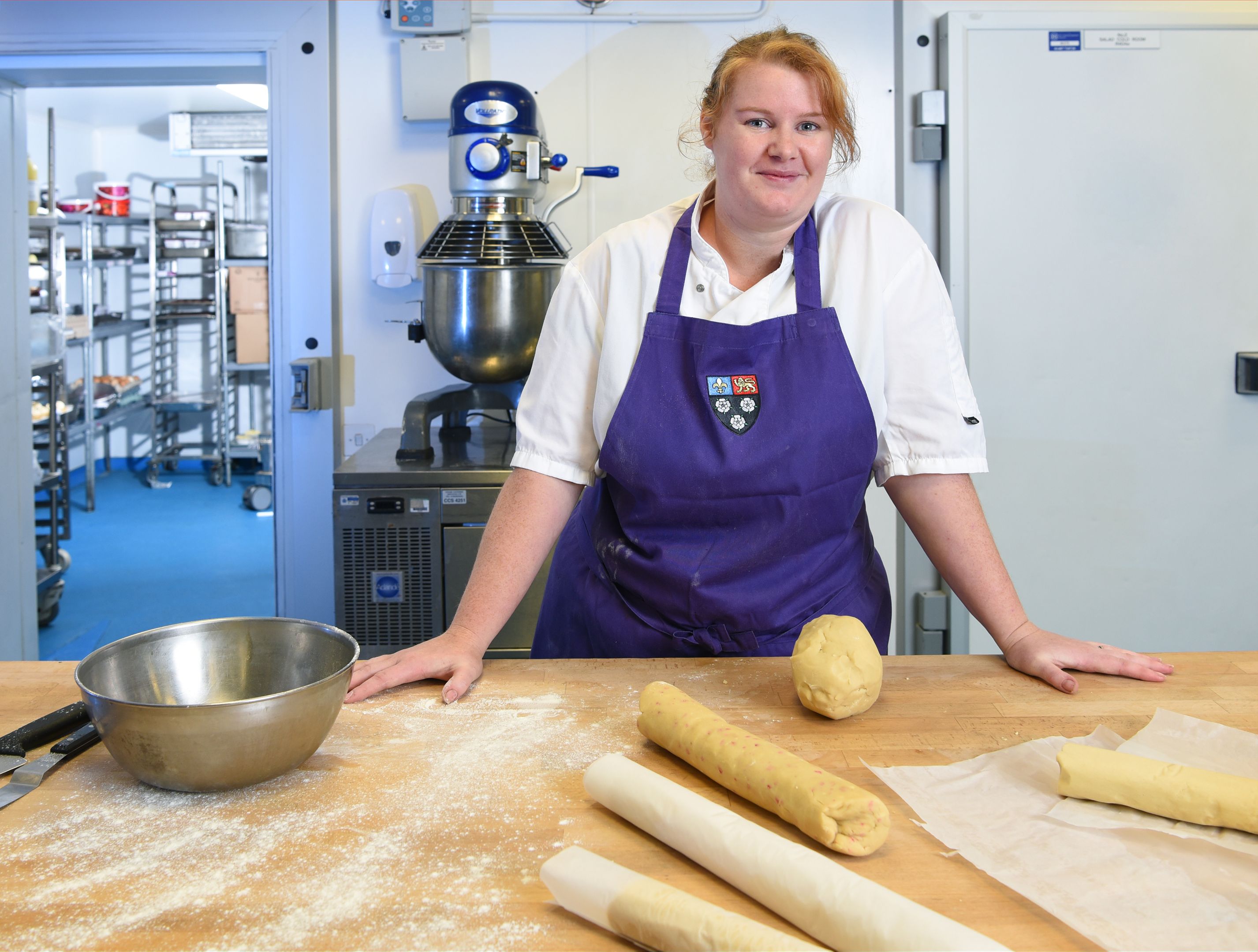 A pastry chef in the College kitchen A woman in a purple apron with dough and baking supplies on a table